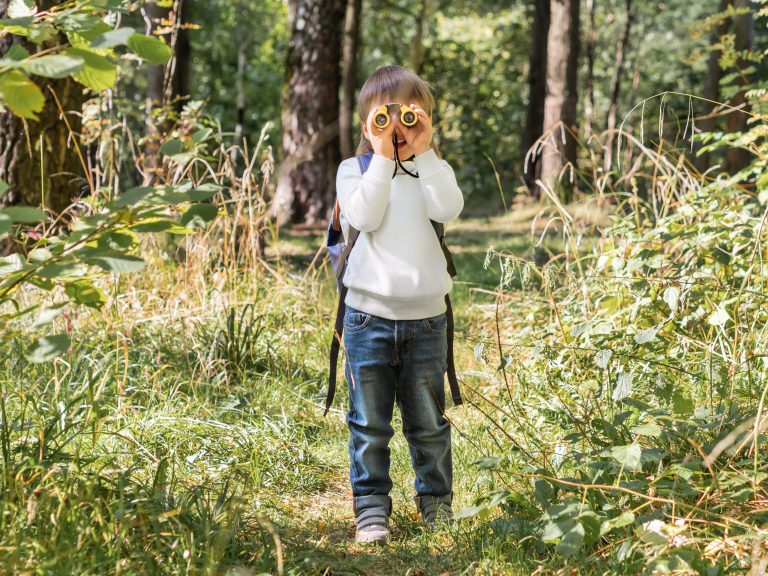 Curious boy is hiking in forest. Outdoor leisure activity for kids. Child looks through binoculars on tree foliage. Sunny day at autumn or summer day.
